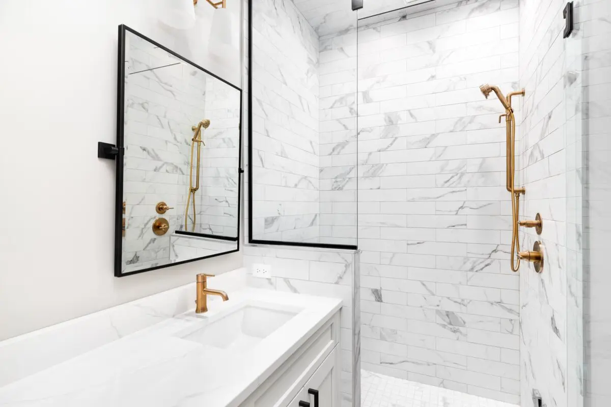 Modern bathroom with white marble tiles, a glass shower enclosure, gold fixtures including faucets and showerhead, a rectangular mirror, and a white vanity with double sinks.