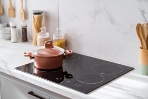 A modern kitchen with a black glass induction cooktop, a covered copper-colored pot on one burner, and jars of pasta, grains, and utensils arranged neatly on a marble countertop and backsplash.
