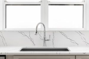 A modern kitchen sink with a curved chrome faucet set in a white marble countertop with gray veining, positioned in front of two large, frosted windows.