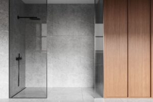 Minimalist bathroom featuring a walk-in shower with gray stone walls and floor, black shower fixtures, and a glass partition next to light wooden cabinets.
