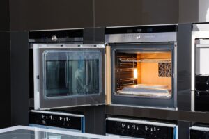 A modern built-in electric oven with its door open, showing empty wire racks and the interior light on. The oven is installed within a glossy black kitchen cabinet.