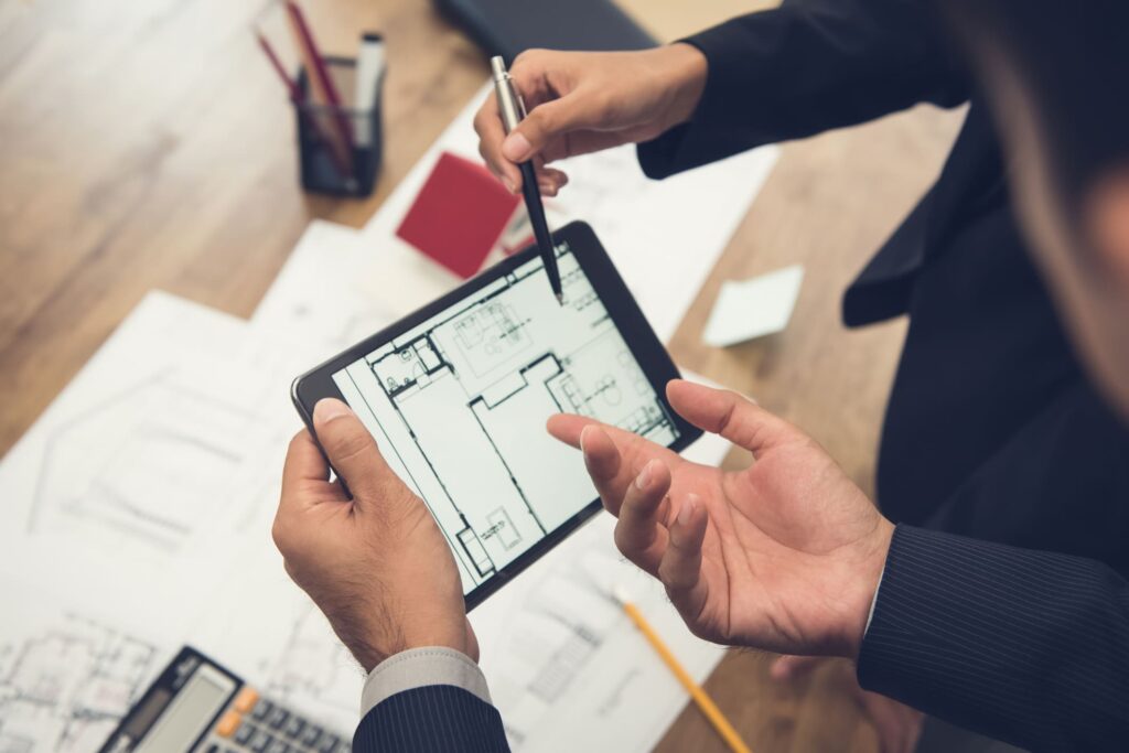 Two people in business attire discuss a commercial renovation in NYC, reviewing a building floor plan on a tablet. Architectural drawings, a calculator, and office supplies are spread across the wooden table.