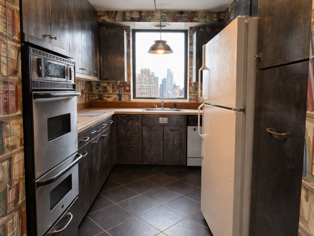 A narrow kitchen with dark cabinets, a white refrigerator, built-in oven, and a window above the sink showing city buildings. The walls are decorated with patterned wallpaper.