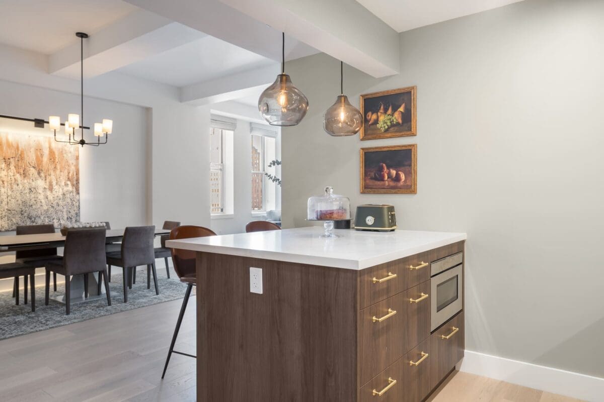 A modern Peninsula Kitchens kitchen with a white countertop island, brown cabinets with gold handles, pendant lights, and wall art; a dining area with a large painting and a chandelier is visible in the background.