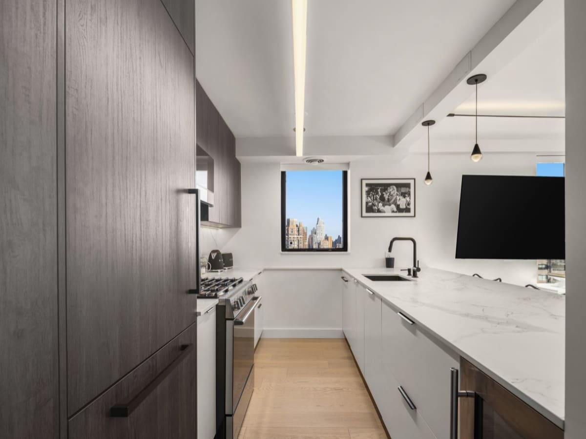 Modern kitchen with dark wood cabinets, white countertops, built-in appliances, and a large window offering a city view. Pendant lights hang above the peninsula, and a framed photo decorates the white wall.