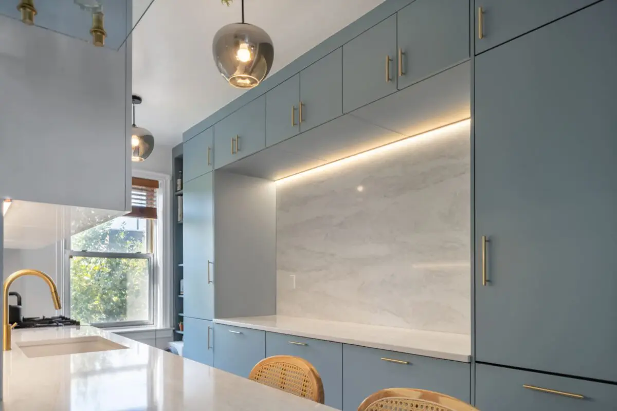 Modern kitchen with light blue cabinets, a marble backsplash, built-in lighting, gold faucet, pendant lights, and natural light coming through a window, with some trees visible outside.