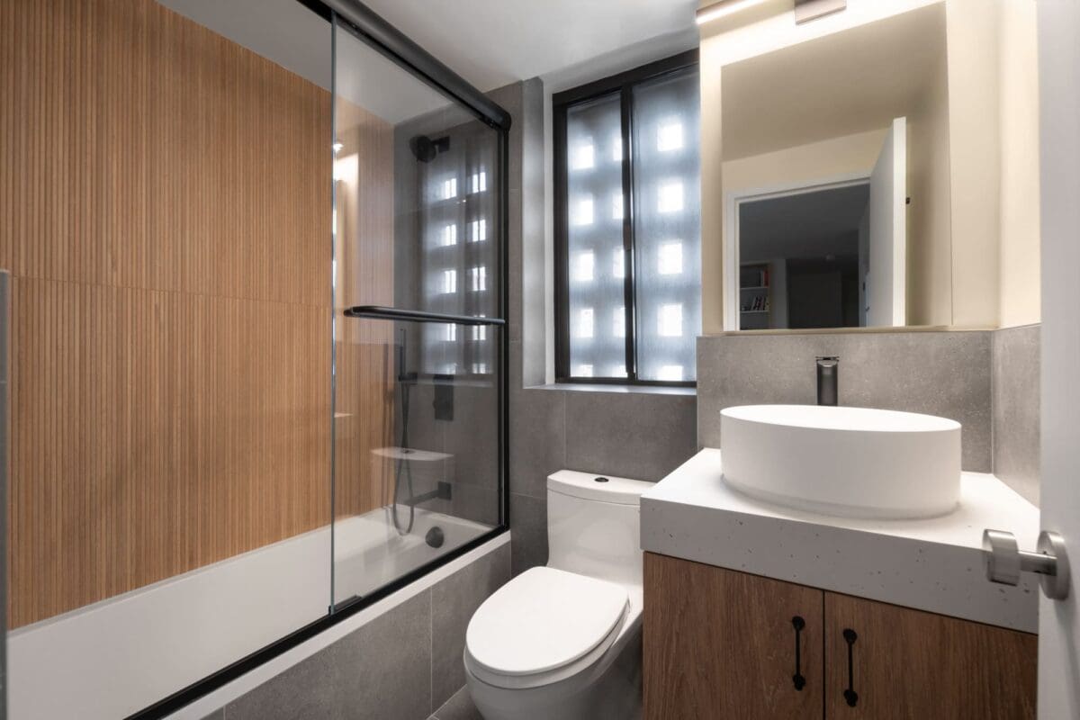 Modern bathroom with wood accent wall in shower, glass shower door, white toilet, round vessel sink on a gray vanity, and frosted windows letting in natural light.