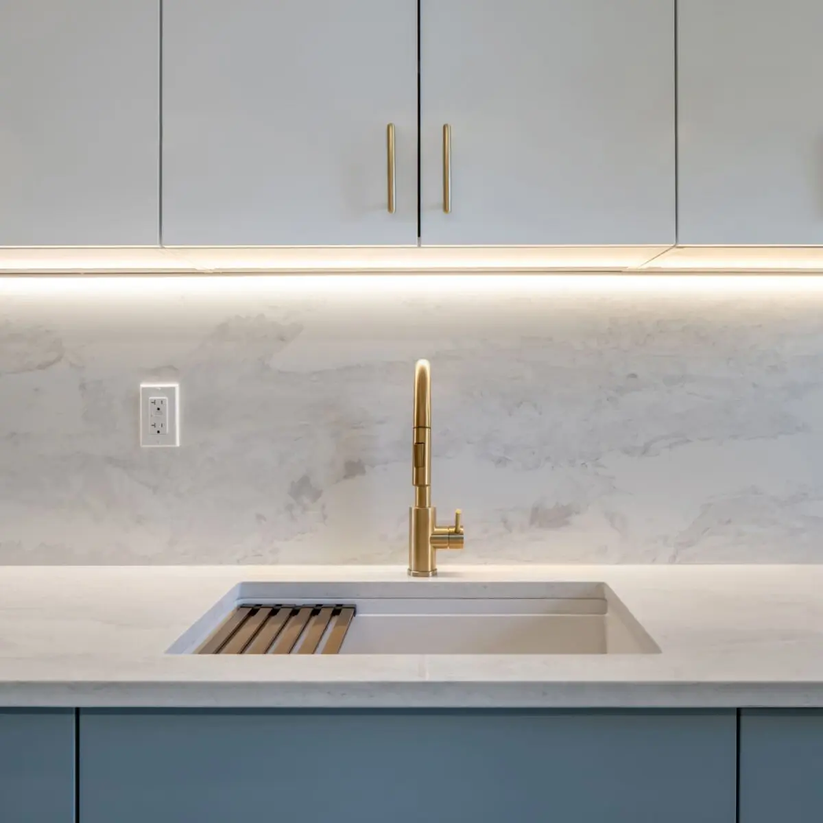 Modern kitchen sink with a gold faucet and marble countertop, set against a marble backsplash. Under-cabinet lighting illuminates the space, and a power outlet is visible on the backsplash to the left of the sink.