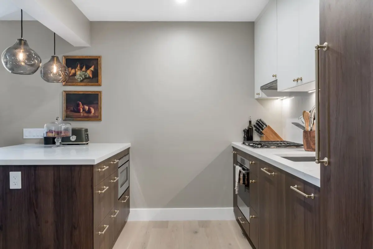 Modern kitchen with dark wood lower cabinets, white upper cabinets, white countertops, and gold hardware. Two pendant lights hang above the counter, and framed artwork is displayed on a neutral gray wall.