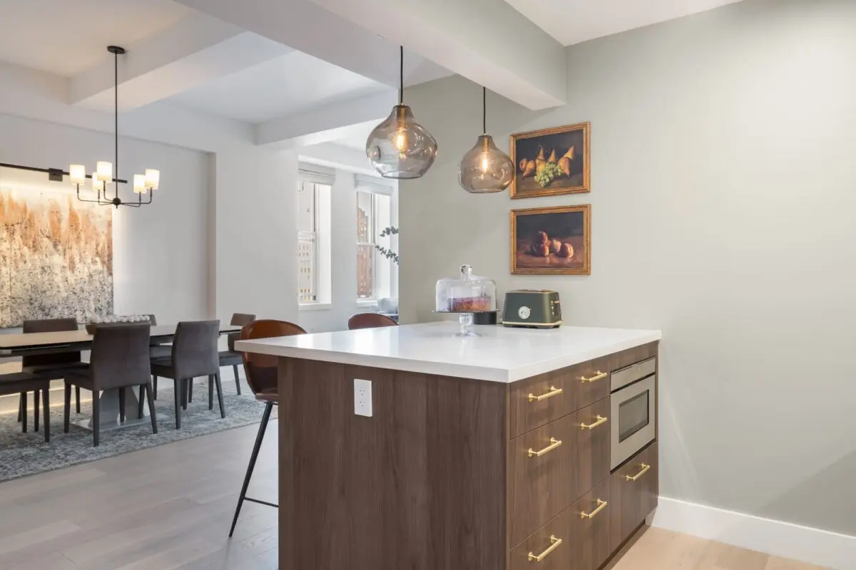 A modern Peninsula Kitchens kitchen with a white countertop island, brown cabinets with gold handles, pendant lights, and wall art; a dining area with a large painting and a chandelier is visible in the background.
