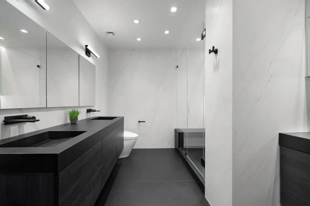 Modern minimalist bathroom with black vanity, dual sinks, large mirror, wall-mounted toilet, glass shower, white marble walls, black floor, and a small potted plant on the counter.