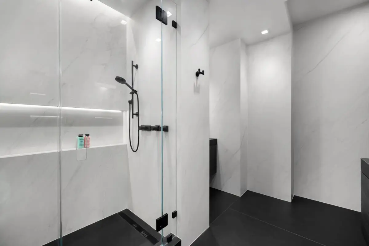Minimalist modern bathroom with a glass shower enclosure, black fixtures, white marble walls, dark flooring, and two bottles on a recessed shower shelf.