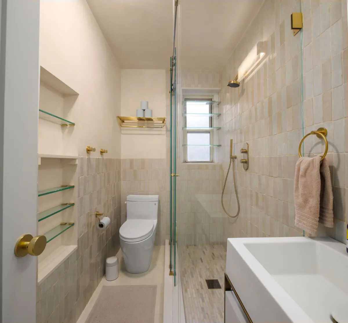 Modern bathroom with beige tiles and gold accents. Features a glass-enclosed shower with rain showerhead, white toilet, wall-mounted shelves, towel rack with beige towel, and a white vanity sink with gold fixtures. A window lets in natural light.