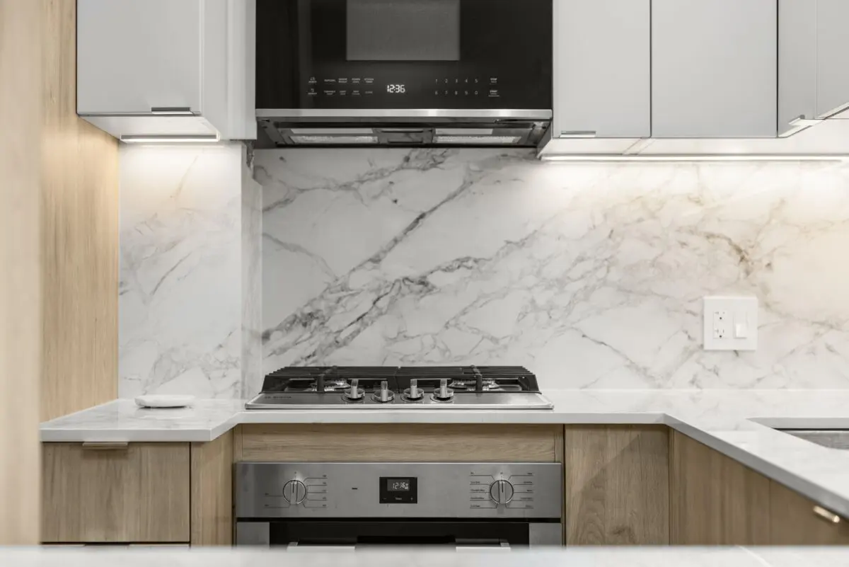 Modern kitchen with a marble backsplash, gas cooktop, built-in oven, white cabinets, wood accents, and under-cabinet lighting. A small item is on the counter near the stovetop.