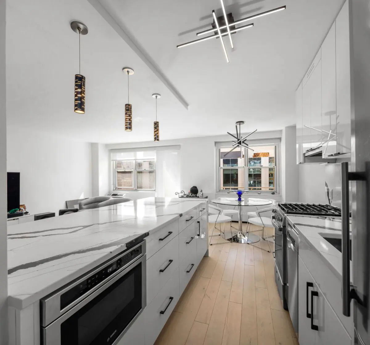 Modern kitchen with a white marble island, built-in oven, and sleek cabinets. Pendant and ceiling lights add elegance. Large windows illuminate the wooden floors. A dining area with a round table and chairs is visible in the background.