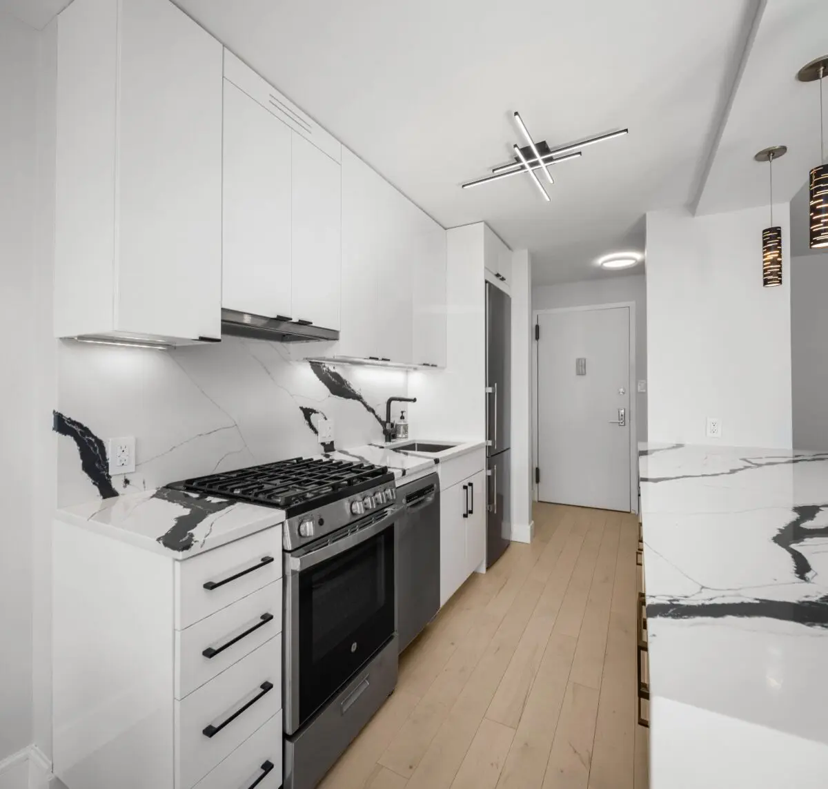 Modern kitchen with white cabinetry, stainless steel appliances, and marble countertops. The stove and oven are built-in, with a fridge beside them. The kitchen has a light wood floor and contemporary overhead lighting.