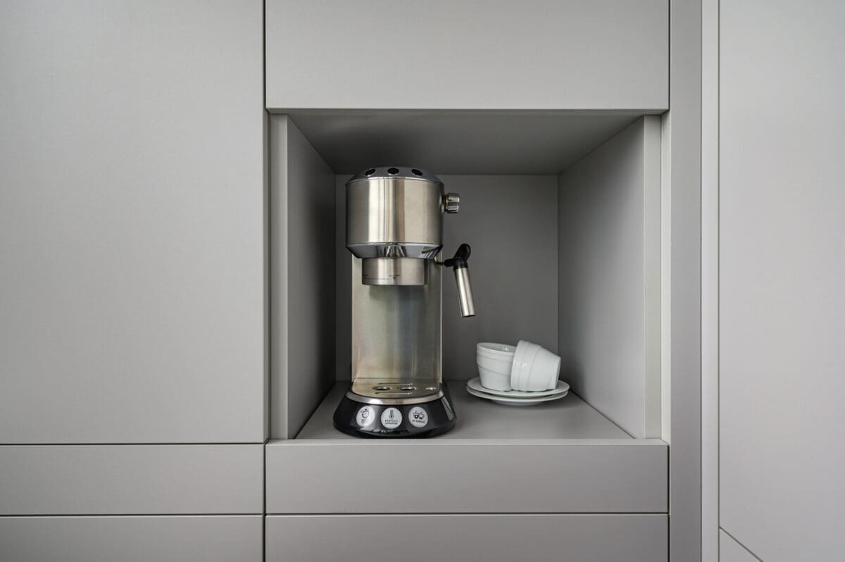 A silver espresso machine sits in a recessed shelf with two stacked white cups and saucers beside it, surrounded by smooth gray cabinetry.