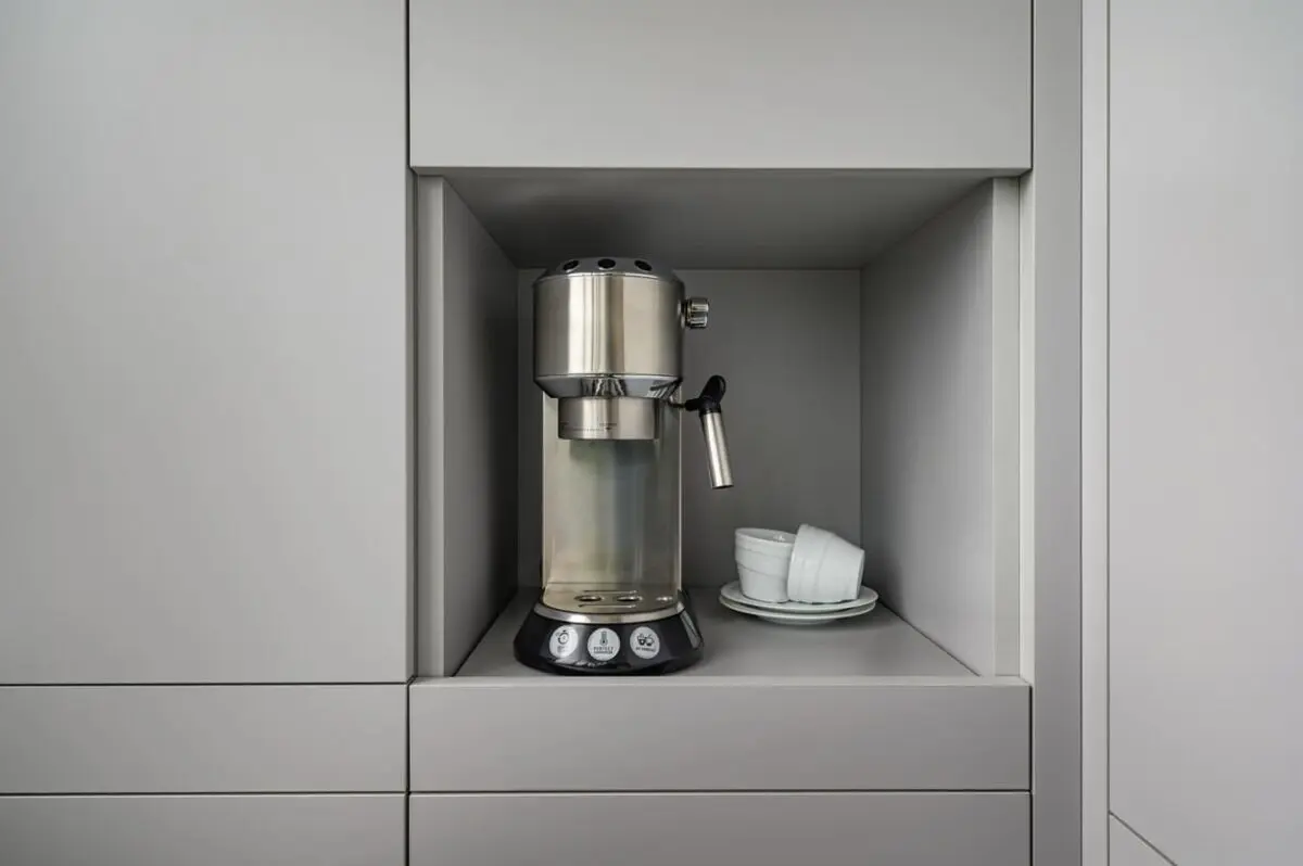 A silver espresso machine sits in a recessed shelf with two stacked white cups and saucers beside it, surrounded by smooth gray cabinetry.