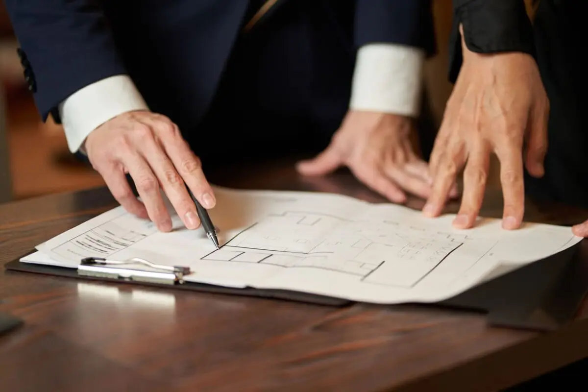 Two people in business attire point at a building blueprint on a clipboard, discussing a restaurant layout and architectural plans on a wooden table.