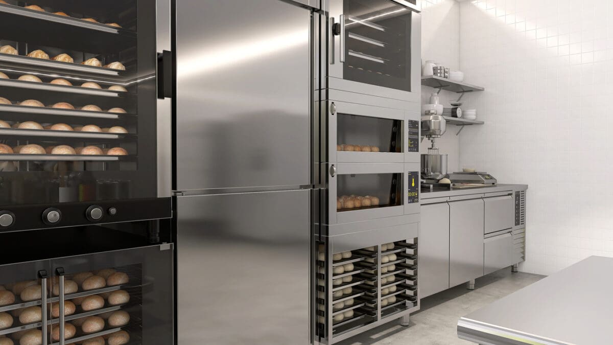 Commercial bakery kitchen with stainless steel appliances, including ovens with bread inside, refrigerators, shelves with equipment, and trays stacked next to a countertop in a clean, well-lit space.