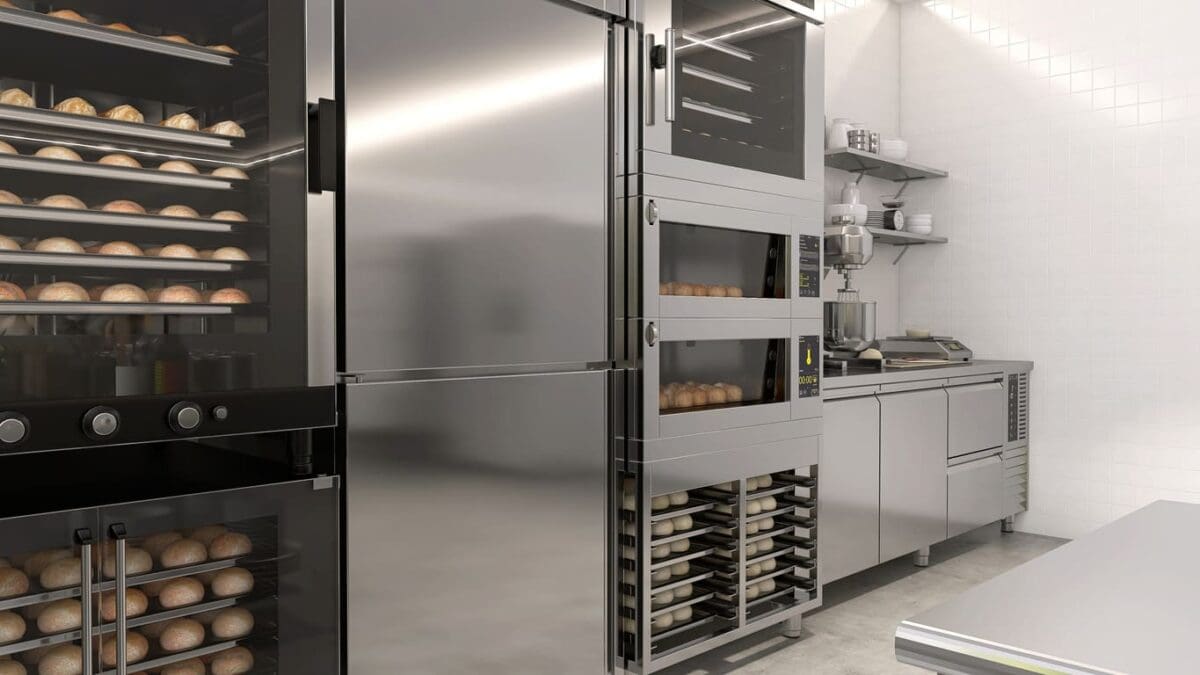 Commercial bakery kitchen with stainless steel appliances, including ovens with bread inside, refrigerators, shelves with equipment, and trays stacked next to a countertop in a clean, well-lit space.