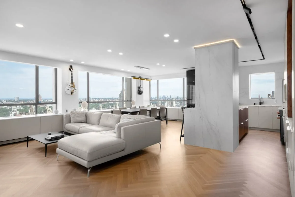 Bright modern living room with large windows, white sectional sofa, coffee table, and open kitchen. Guitars hang on the wall. City skyline visible through the windows. Wood herringbone floor throughout the space.