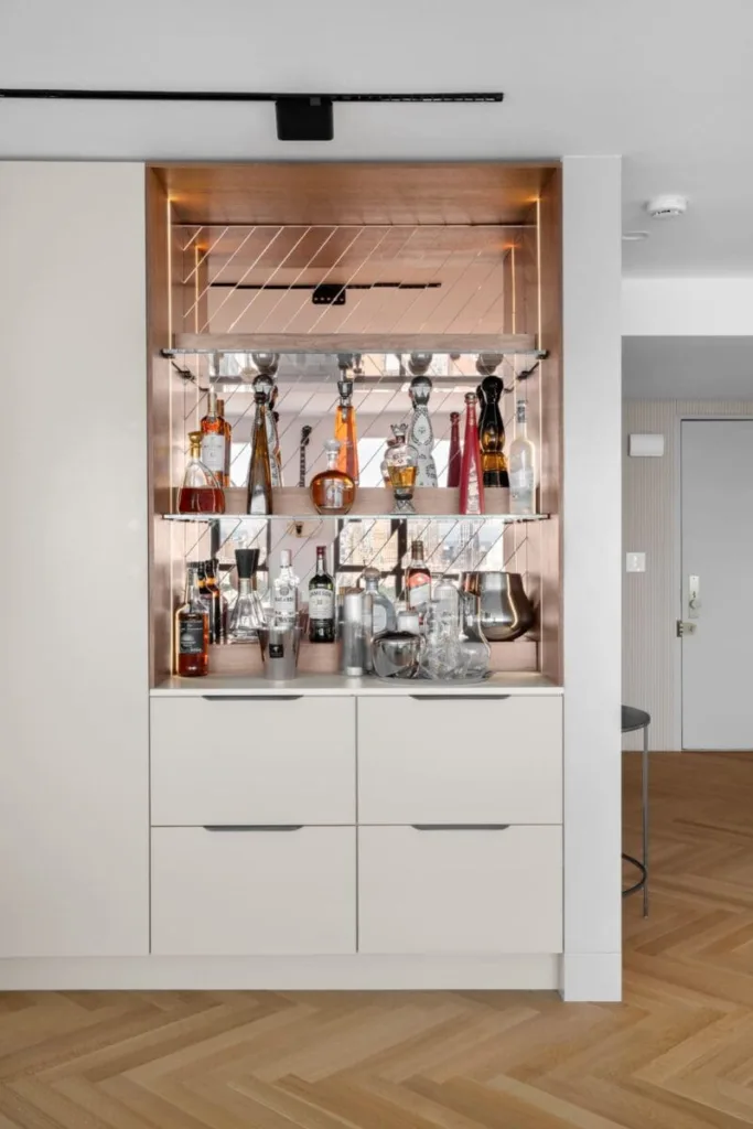 A modern home bar with a copper backsplash, shelves of liquor bottles, glasses, and bar tools above white cabinets. The setup is neat and stylish, blending into a bright, contemporary room with wood flooring.