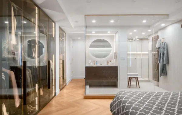 Modern bedroom with a glass-walled bathroom, circular mirror, walk-in closet with glass doors, wood herringbone floors, and gray patterned bedding in the foreground.