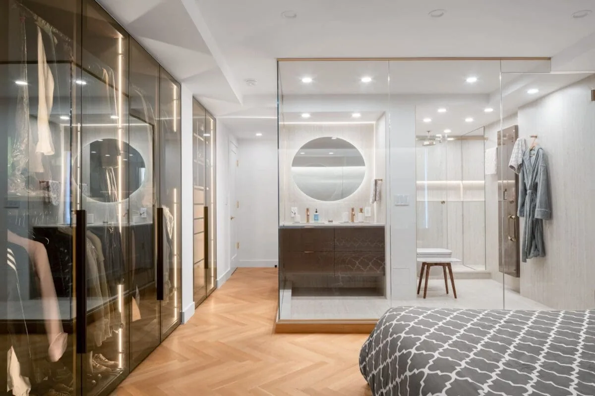 Modern bedroom with a glass-walled bathroom, circular mirror, walk-in closet with glass doors, wood herringbone floors, and gray patterned bedding in the foreground.