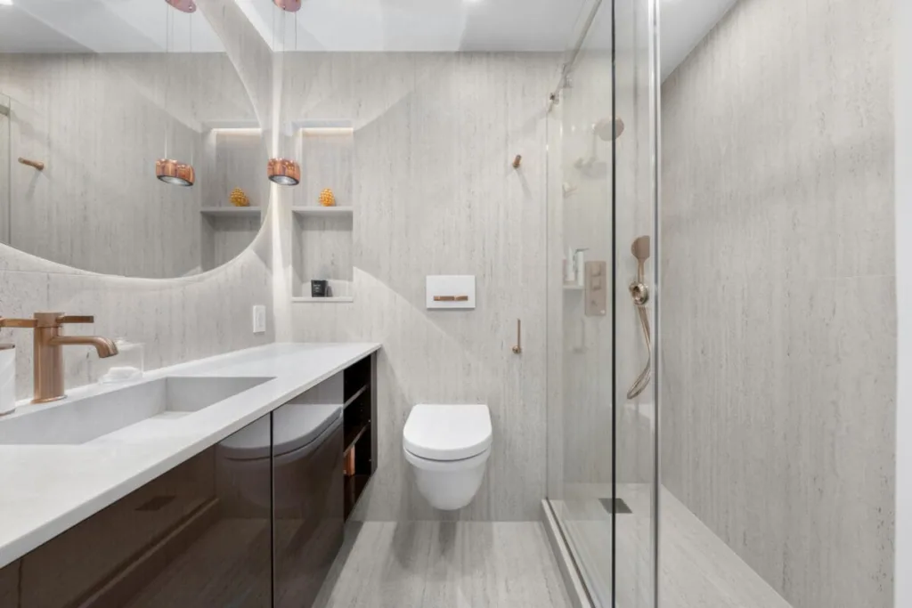 Modern bathroom with floating toilet, double sink vanity, glass shower, large mirror, and built-in shelves holding small decor items. Light neutral tones and minimalist design create a clean, spacious look.