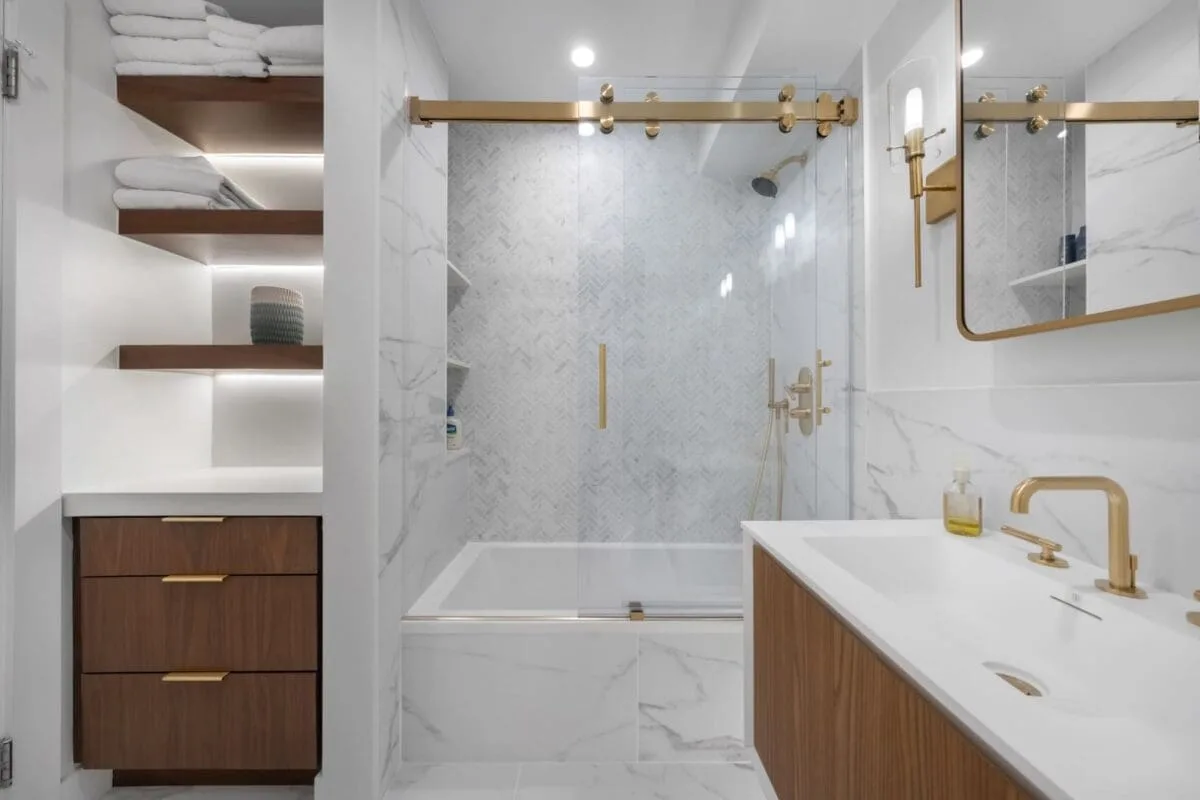 Modern bathroom with white marble surfaces, wooden shelves and drawers, gold fixtures, a bathtub with a glass door, and neatly stacked towels on shelves. The space is well-lit and organized.