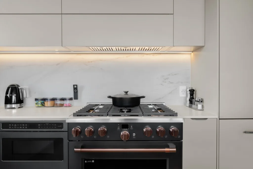 Black gas range with copper-toned knobs beneath a concealed hood, light cabinetry, and an illuminated veined backsplash.