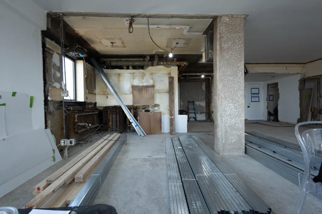 Kitchen during demolition, with partition walls removed, exposed ceiling pipes, and a concrete column beside the living area.