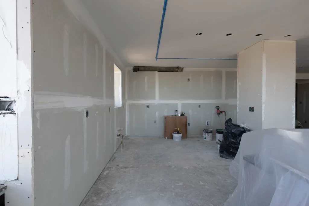 Kitchen renovation with newly installed drywall, taped wall seams, ceiling light openings, and exposed plumbing connections.